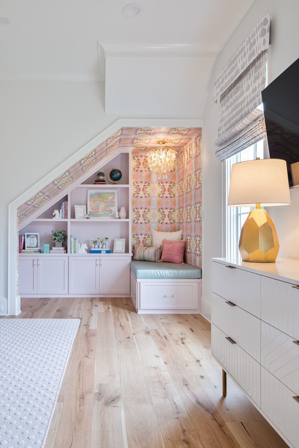 Built-in pink shelving and a cozy window seat with patterned wallpaper and a gold lamp on a white dresser in a room.