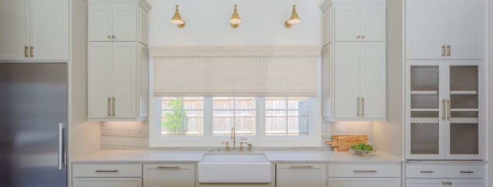 A bright, modern kitchen featuring white cabinets, a farmhouse sink under a window with a striped shade, and gold accents.