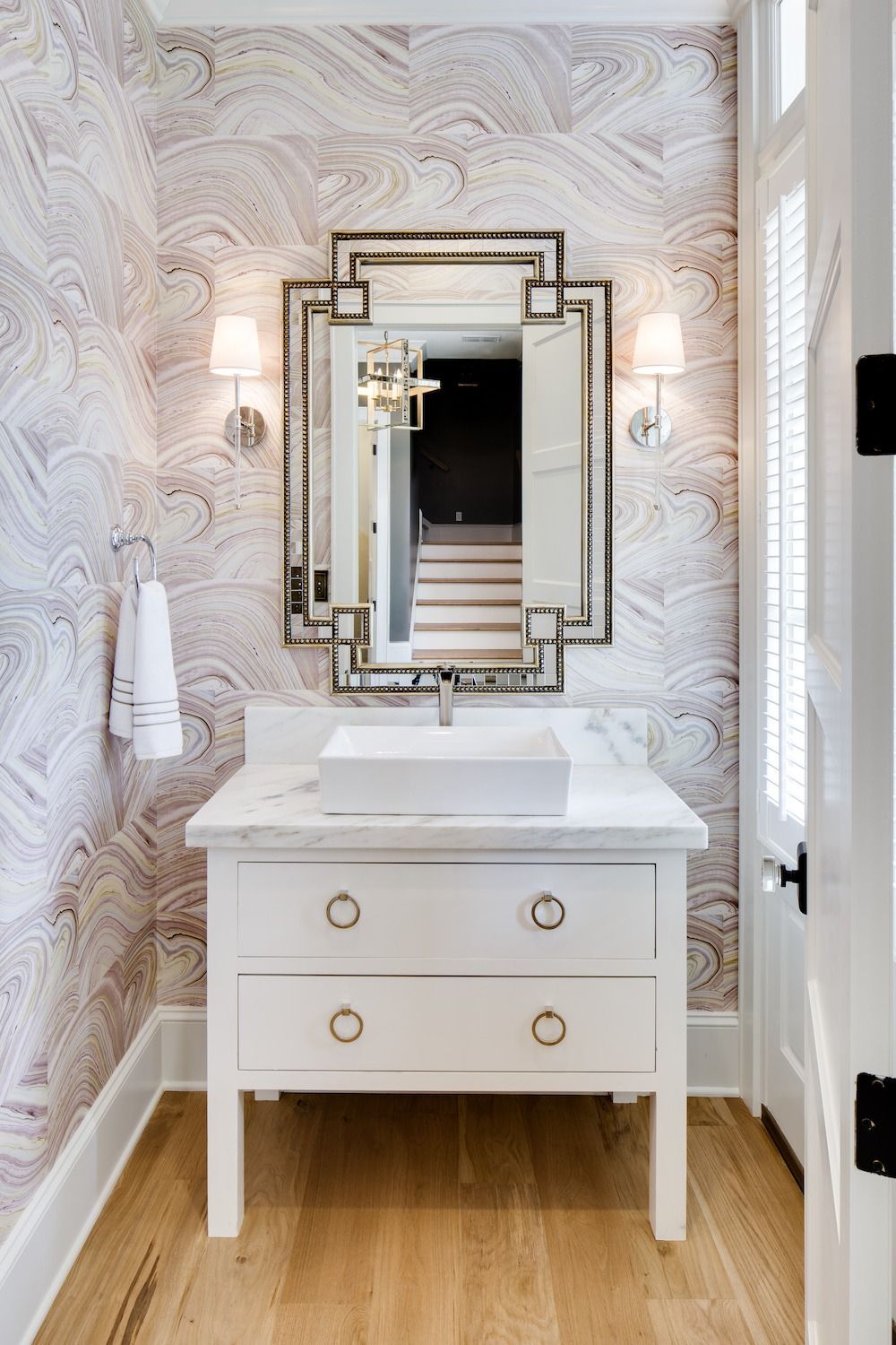 A white vanity with a rectangular sink, ornate mirror, and wall sconces against swirling patterned wallpaper in a bathroom.