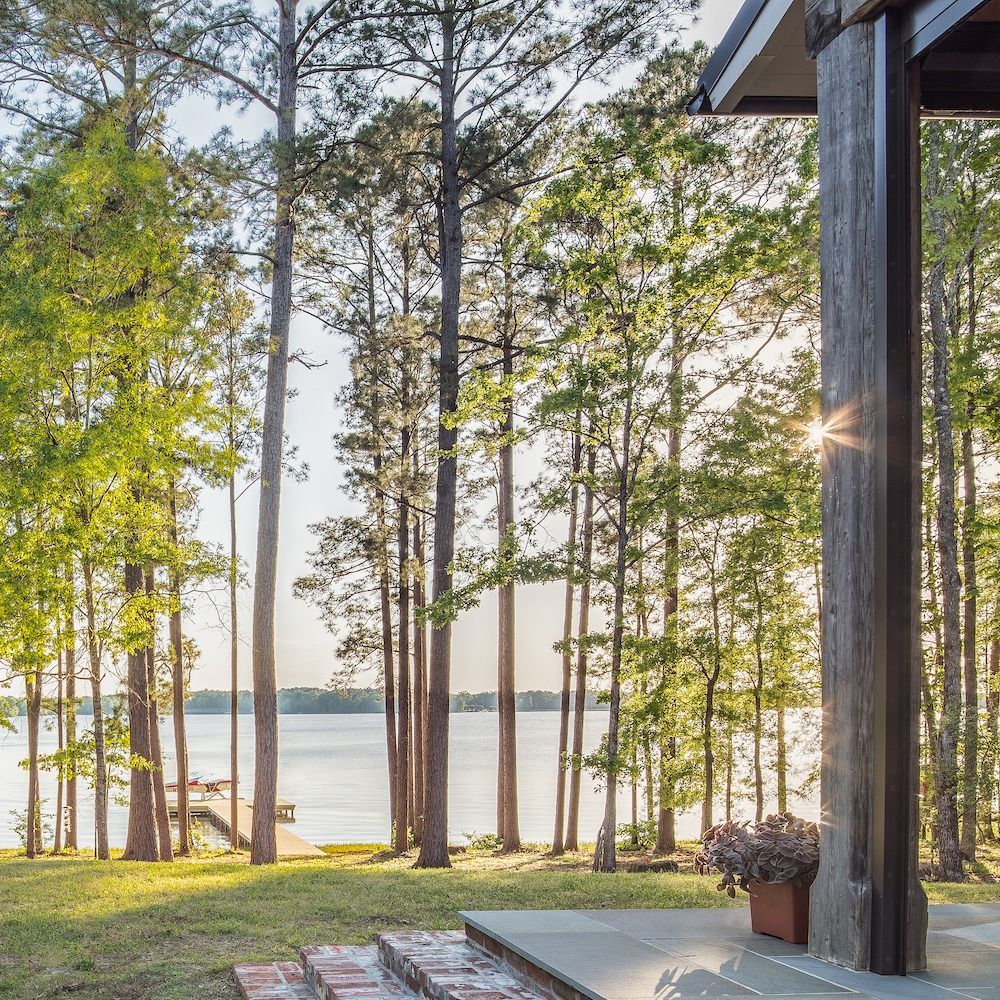 A wooden post on a patio overlooks a grassy lawn, trees, and a lake with a small dock in the distance under bright sunlight.
