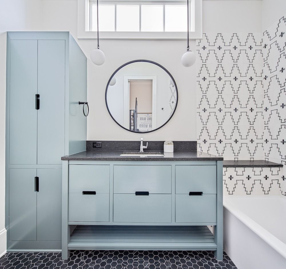 A modern bathroom with light blue cabinets, a round mirror, black hardware, and patterned black-and-white tile work.