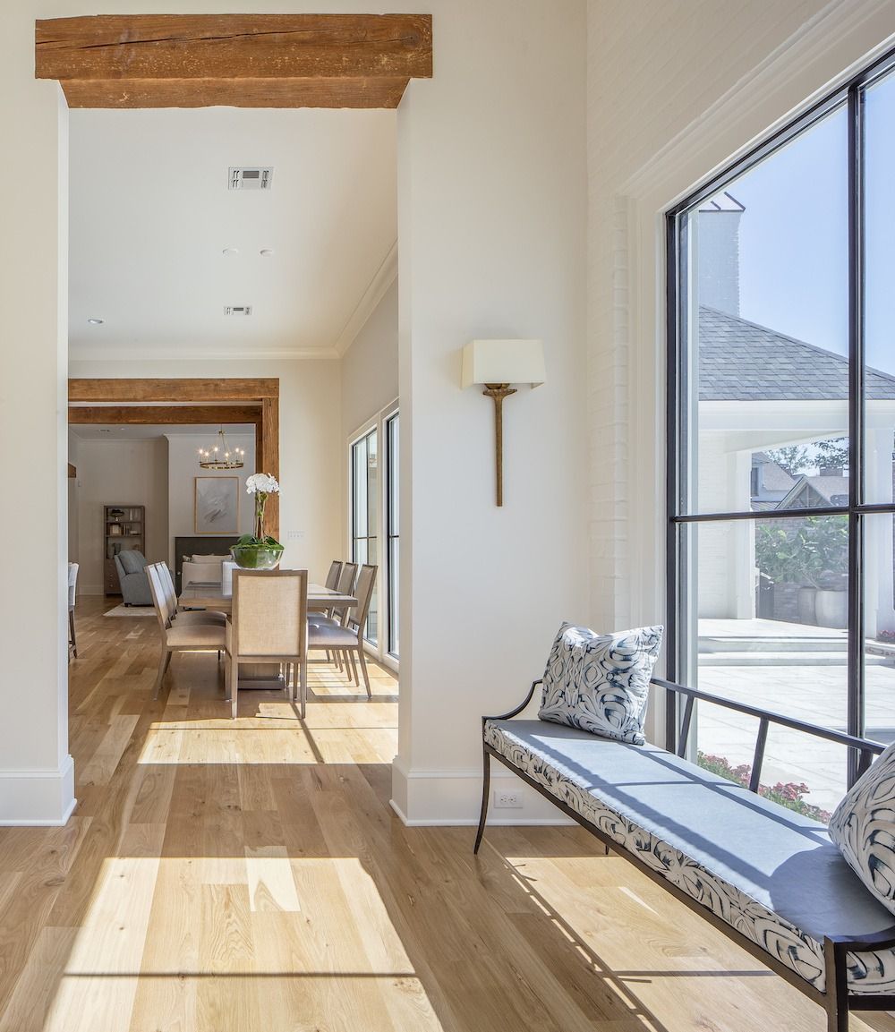 A sunlit hallway with hardwood floors, a metal bench by a large window, and a view into a dining area under wooden beams.