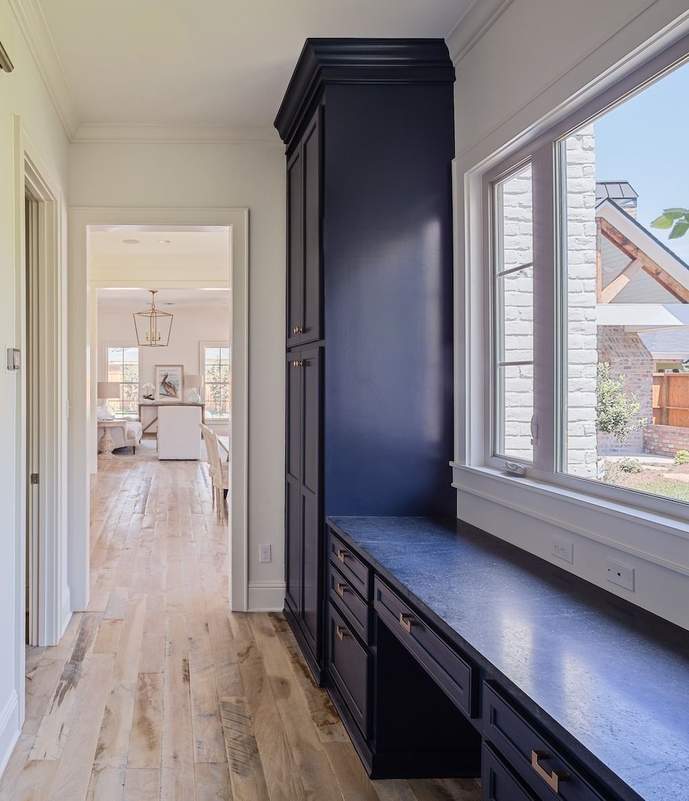 A hallway with dark navy built-in cabinets and a desk, light wood floors, and a window looking out onto a patio.