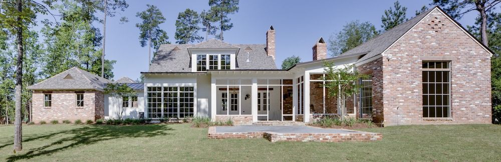 A multi-sectioned brick house with large windows and chimneys sits on a green lawn against a clear blue sky.