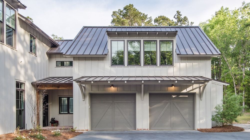 A two-story house exterior with white board-and-batten siding, a dark metal roof, and two garage doors.