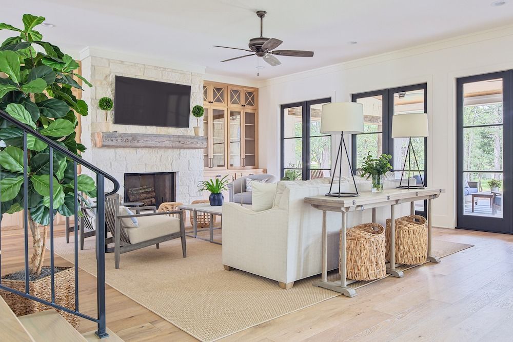 A bright, airy living room with a fireplace, white sofa, neutral area rug, and large windows looking out to greenery.
