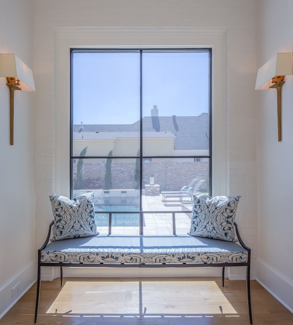 A metal bench with blue patterned cushions sits in a nook between two wall sconces in front of a large window.