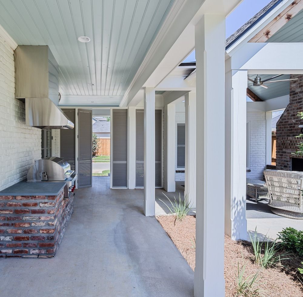 A covered outdoor kitchen area with a brick counter, stainless steel grill, light blue ceiling, and white support columns.