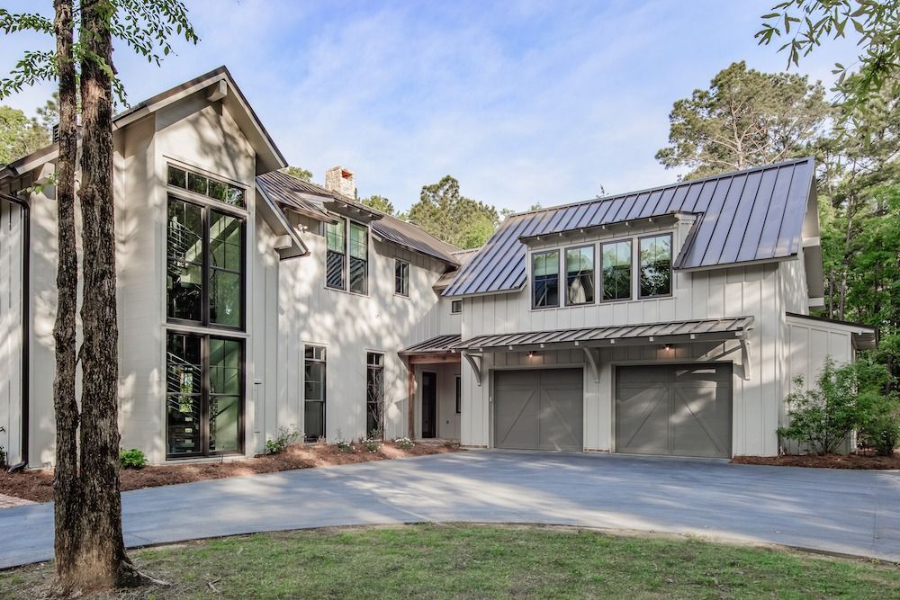 A two-story light-colored house with tall windows, a metal roof, and an attached two-car garage set in a wooded area.