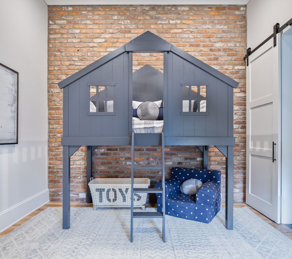 A dark gray house-shaped loft bed stands against an exposed brick wall, with a toy bin and blue chair underneath.