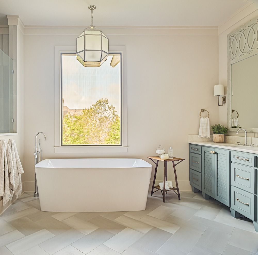 A bright, modern bathroom featuring a white freestanding tub, light gray herringbone tile, and blue-gray cabinetry.