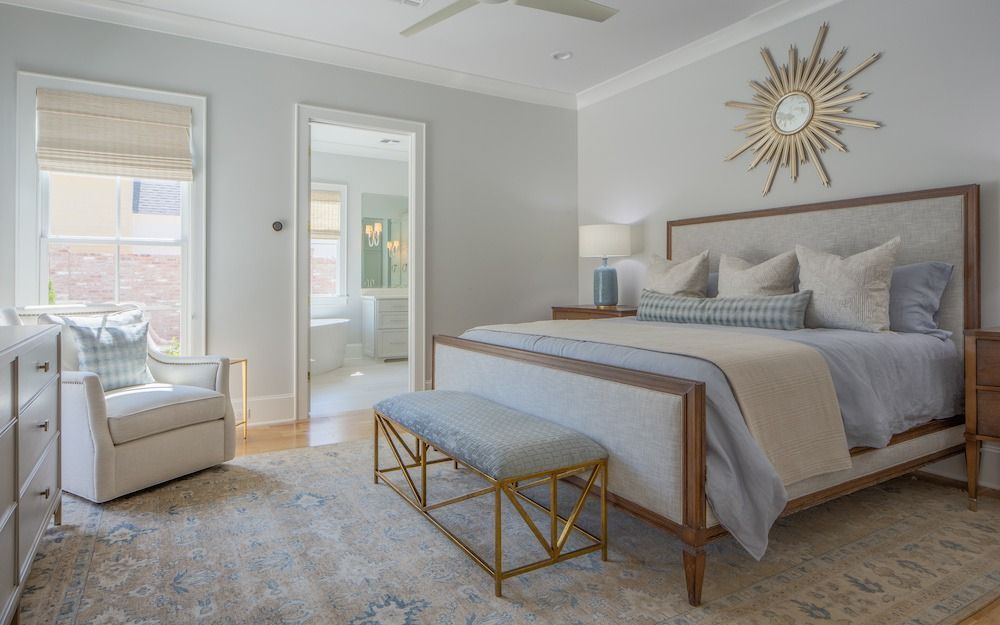 A bright bedroom featuring a queen bed with a tufted headboard, a gold-based bench, an armchair, and a sunburst wall mirror.