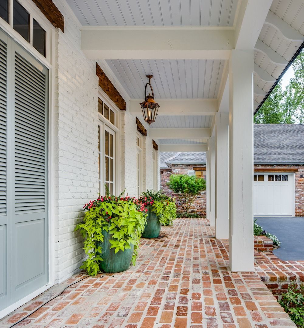 A covered porch with white brick walls, a red brick floor, and green potted plants, featuring a hanging lantern light.