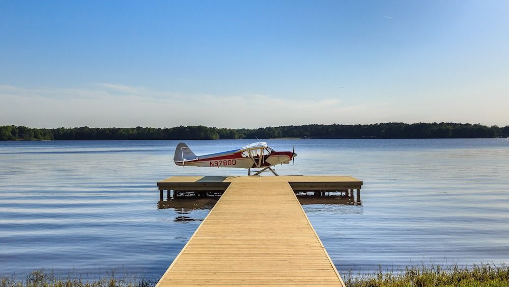 A small red and white floatplane sits parked at the end of a wooden dock extending into a calm lake under a clear blue sky.
