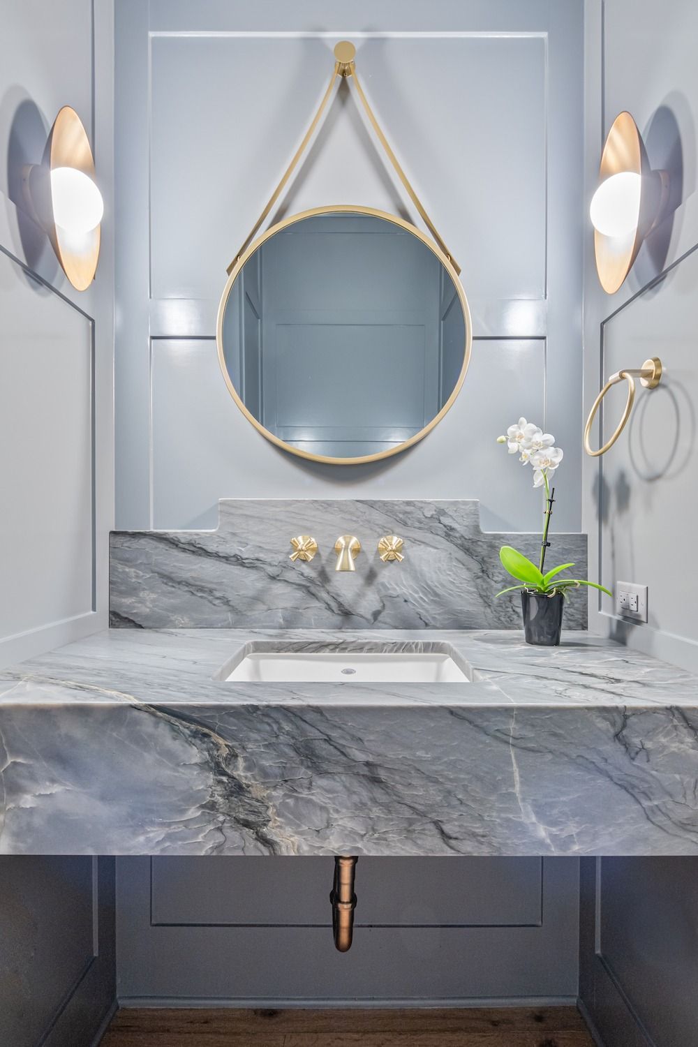 Modern bathroom vanity with grey marble countertop, a circular hanging mirror, gold fixtures, and wall sconces.