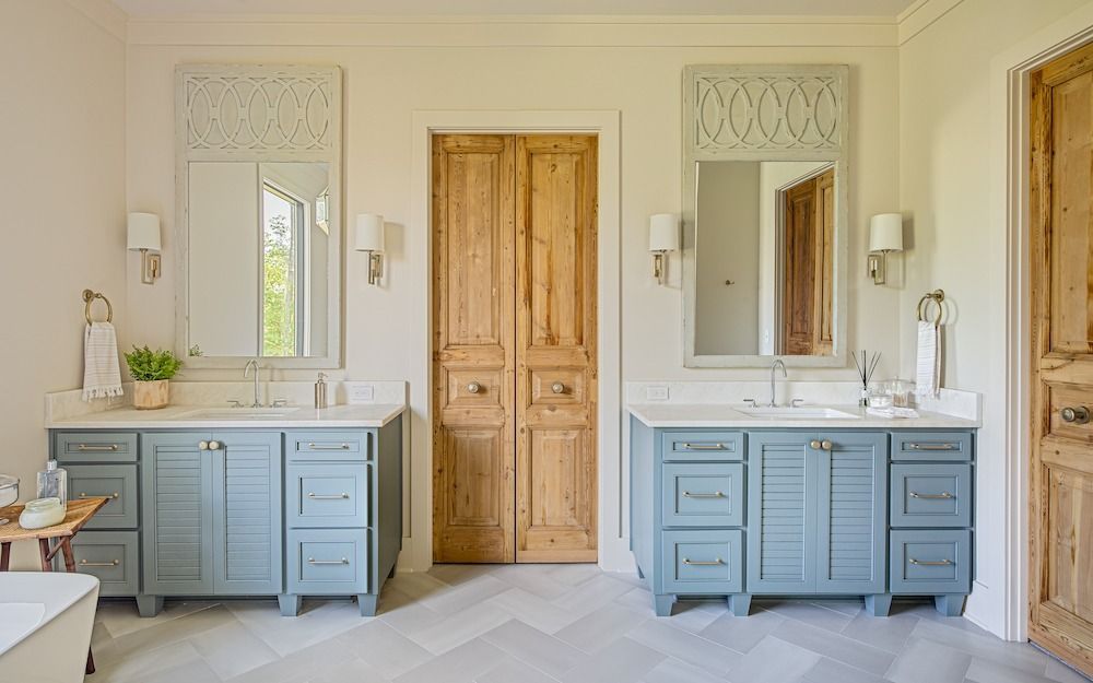 A bright bathroom with two blue shuttered vanity cabinets, white countertops, matching mirrors, and a wooden door.