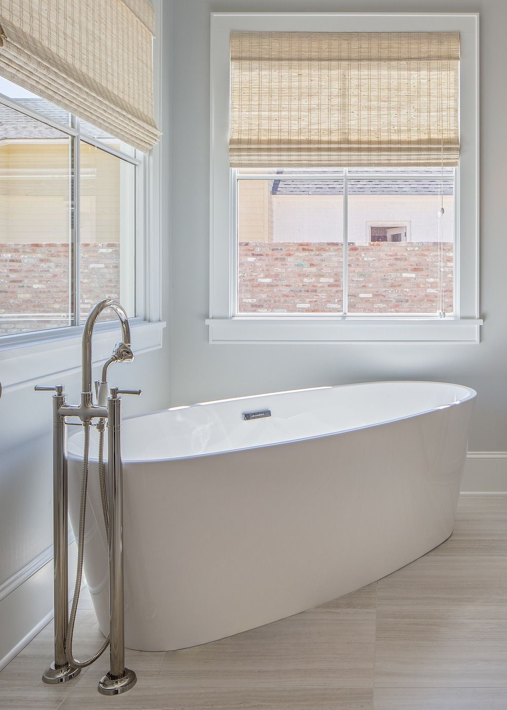 A white freestanding bathtub stands in a bathroom corner near a faucet and two windows with light-colored bamboo shades.