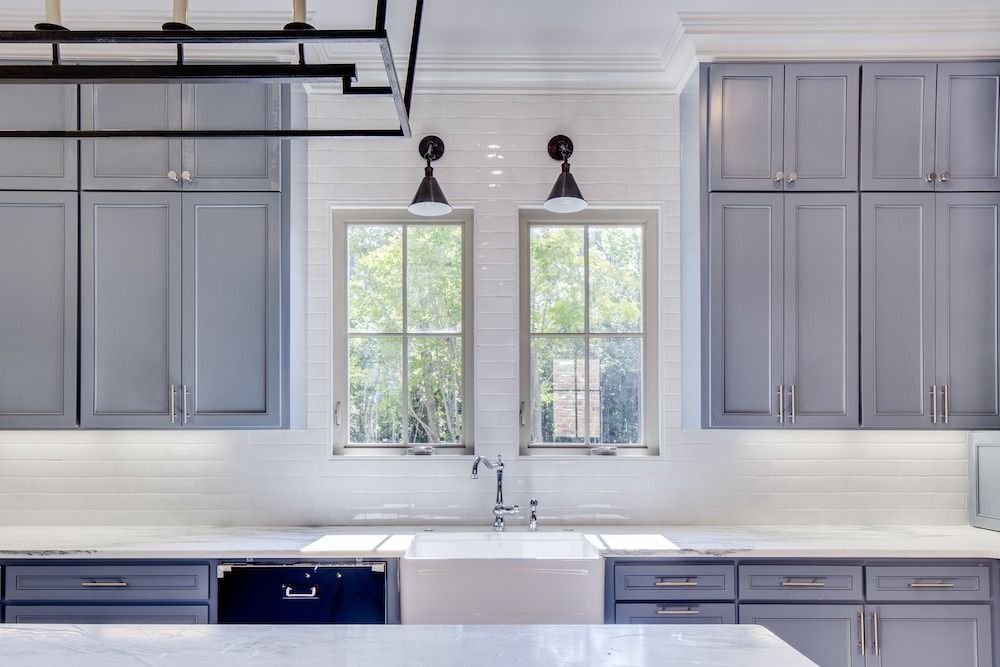 Kitchen view with blue-grey cabinets, white tile backsplash, a farmhouse sink under two windows, and black wall sconces.