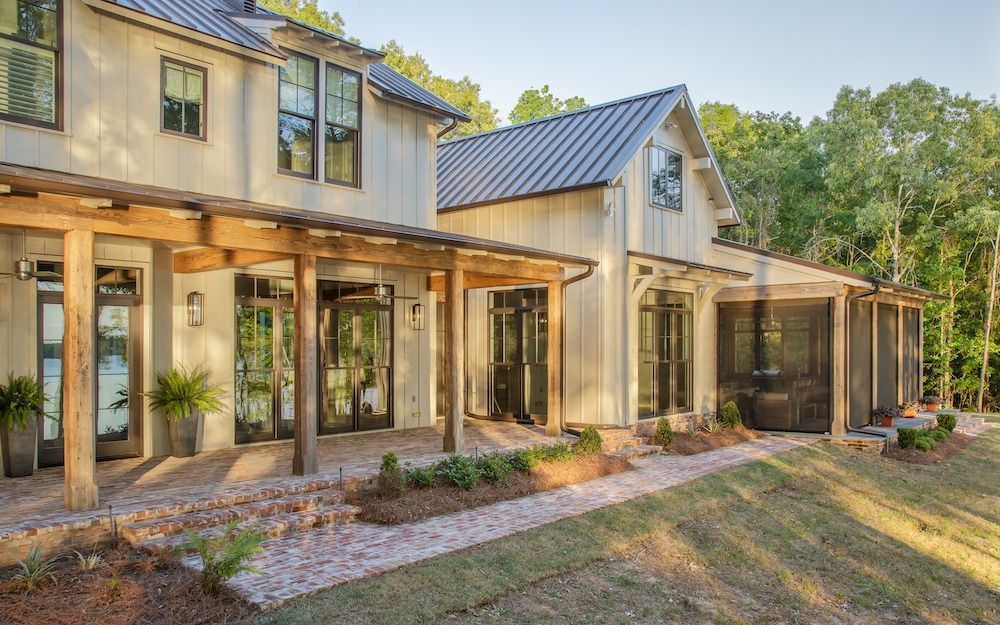 A modern farmhouse with light siding, a dark metal roof, and a wood-beamed porch overlooking a brick patio and lawn.