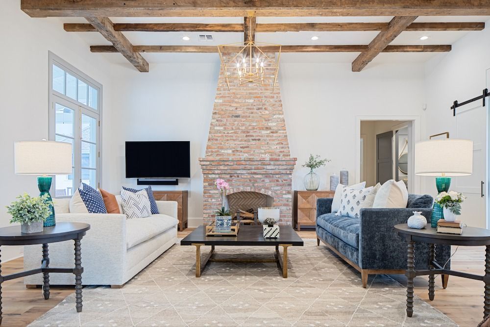 A bright living room with exposed wood ceiling beams, a central stone fireplace, a white sofa, and a dark blue sofa.
