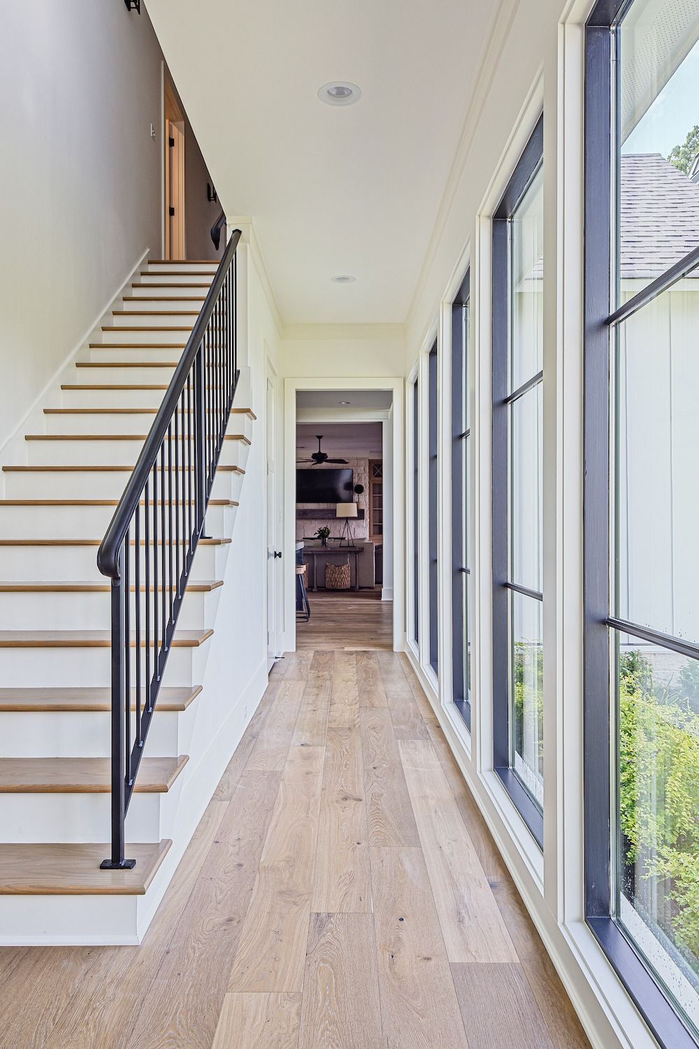A bright hallway with light wood flooring, a staircase with black metal railings, and floor-to-ceiling windows on the right.