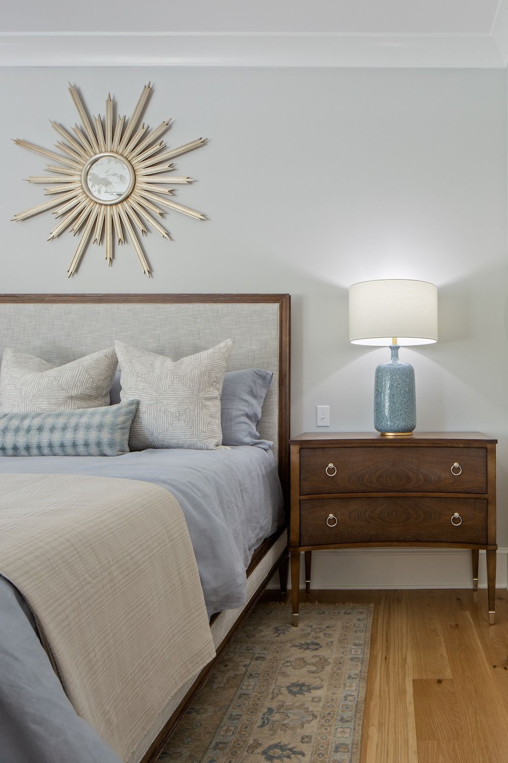 A cozy bedroom scene featuring a bed with gray bedding, a wooden nightstand with a blue lamp, and a gold sunburst mirror.