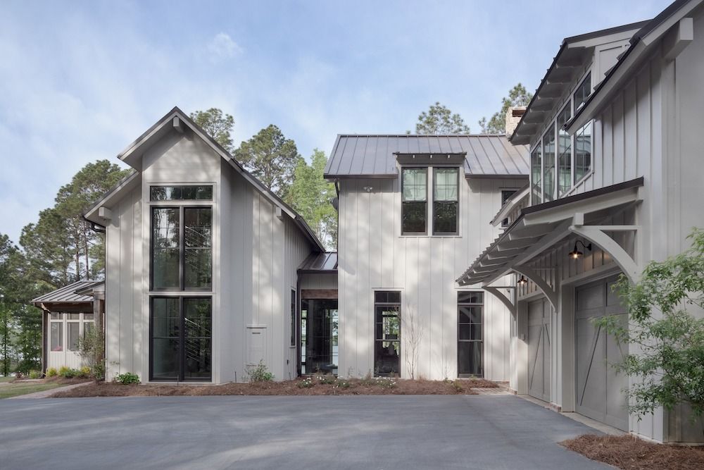 A modern, off-white house with a metal roof and large glass windows, nestled among trees next to a paved driveway.