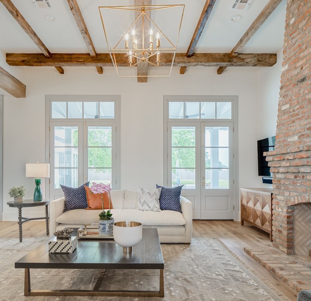 A bright, modern living room featuring a white sofa, dark coffee table, rustic brick fireplace, and exposed wood beams.