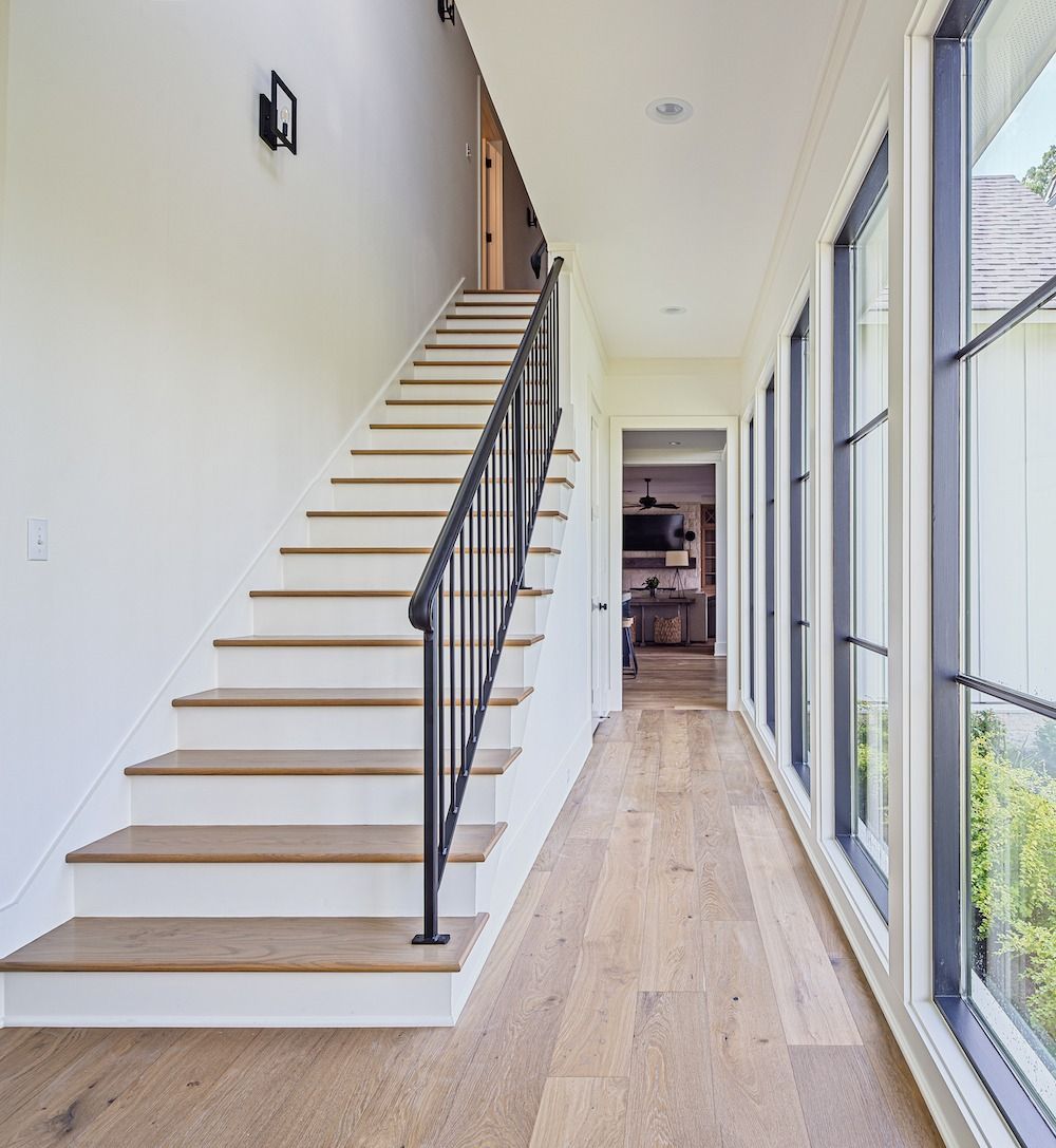 A bright hallway with light wood floors, a staircase with black metal railings, and a wall of floor-to-ceiling windows.