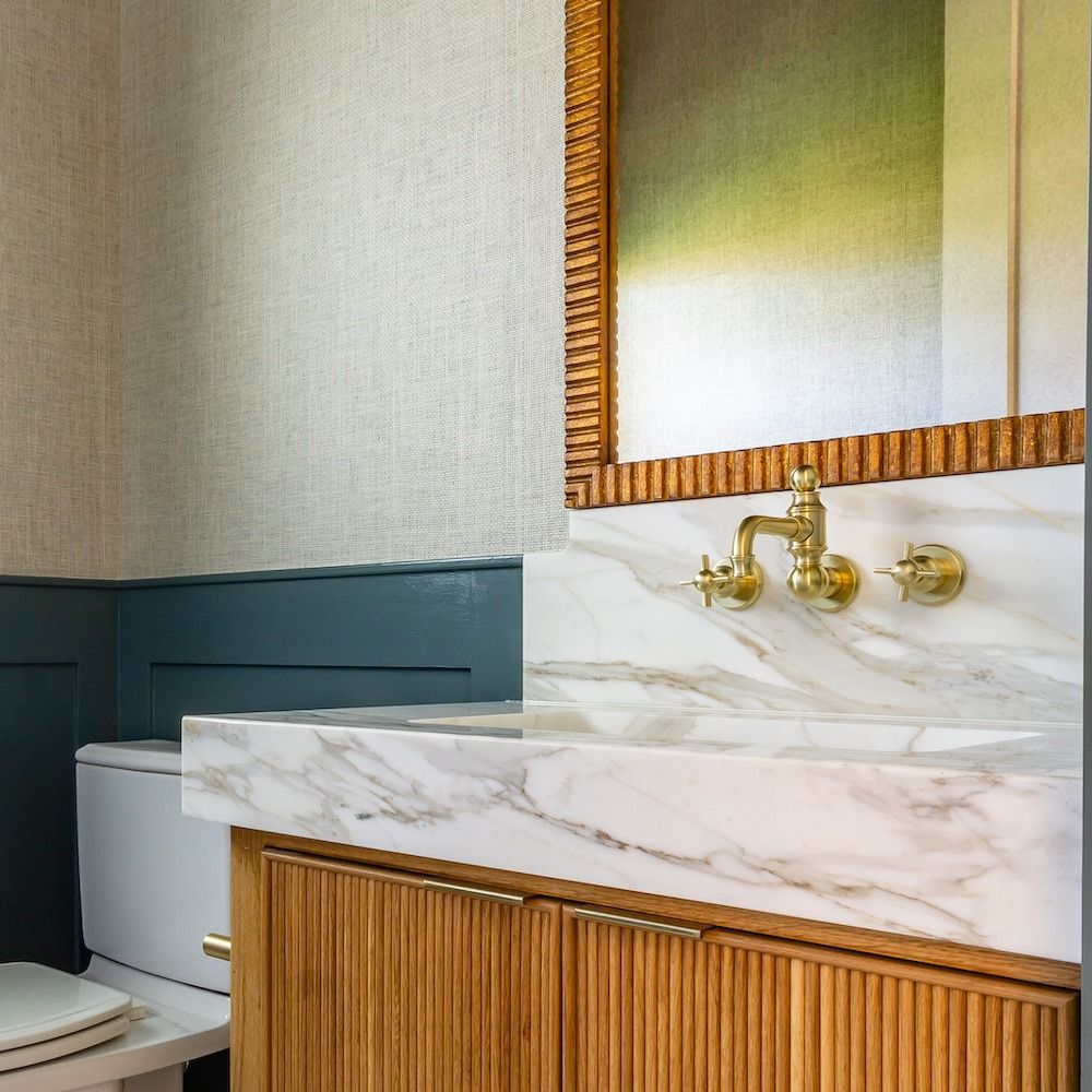 A bathroom vanity with a marble sink, gold wall-mounted faucet, reeded wood cabinet, and a textured gold-framed mirror.