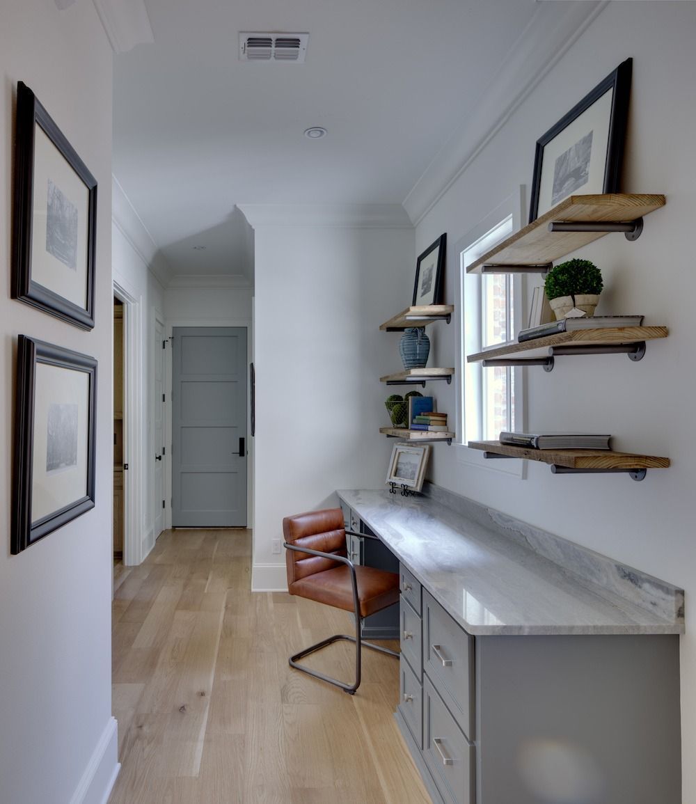 A hallway office nook with a gray built-in desk, leather chair, wood shelves, and framed art on white walls.