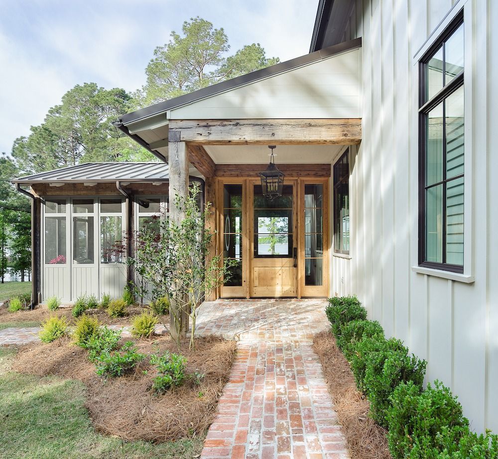 A modern farmhouse entrance with white vertical siding, a brick walkway, and a wooden front door, set beside pine trees.
