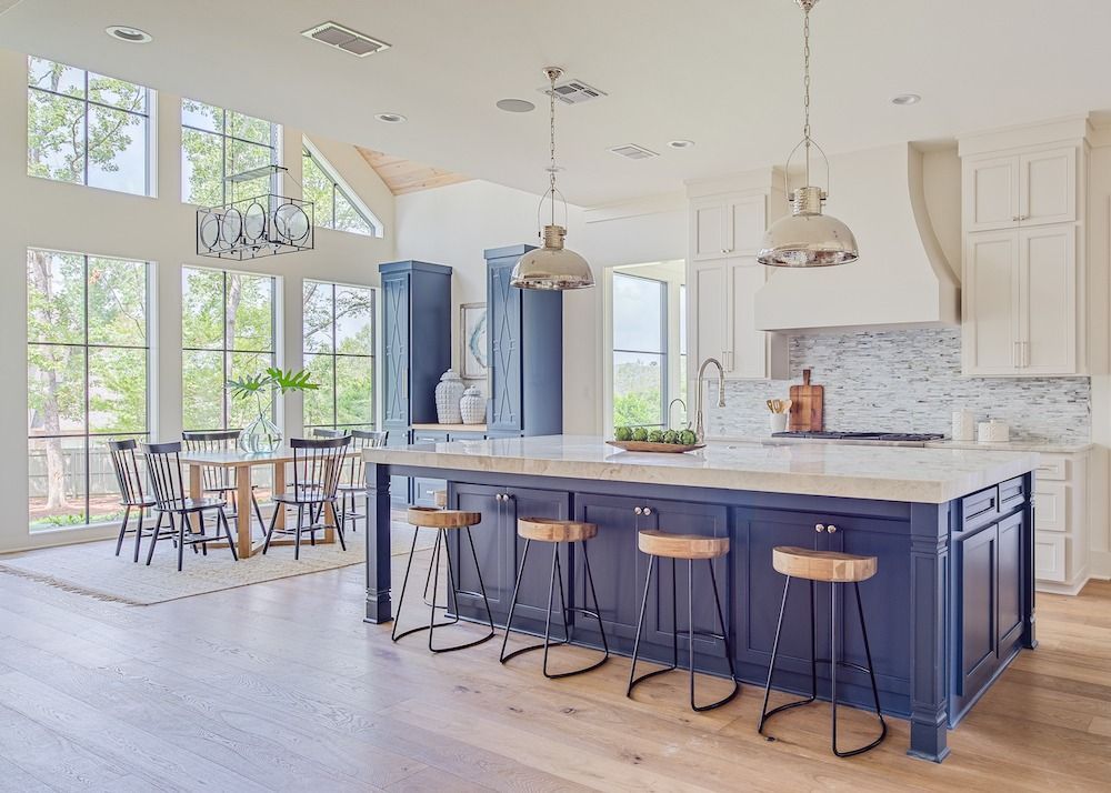 A modern kitchen with a large blue island, light wood floors, white cabinets, and a dining area beside tall windows.