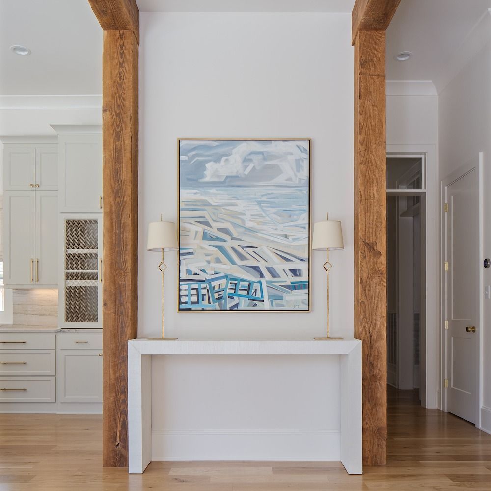 A white console table sits between two rustic wooden columns, topped by two gold lamps and a coastal abstract painting.