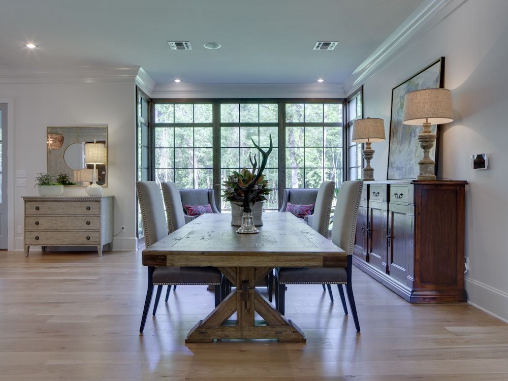 A bright dining room with a wooden table, six upholstered chairs, a sideboard with two lamps, and large window wall.