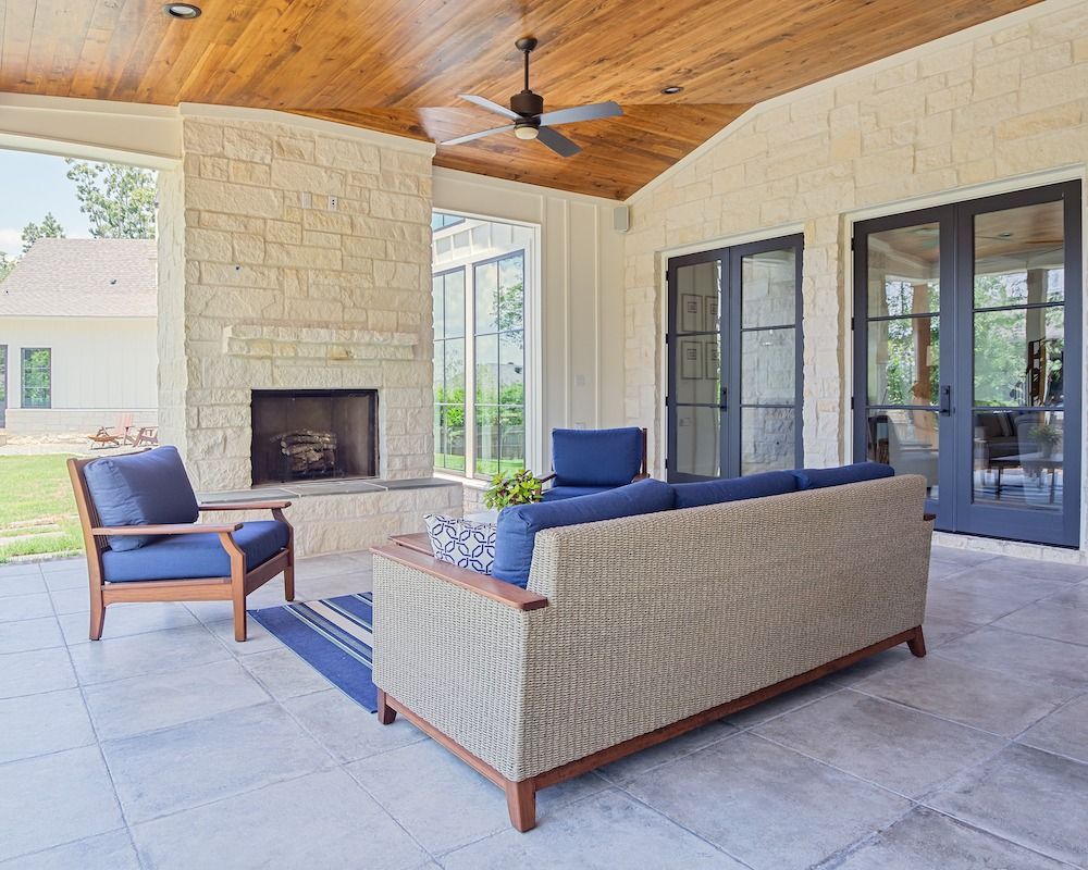 A stone-walled patio with a fireplace, wicker sofa, blue armchair, wood ceiling, and glass doors leading to the house.