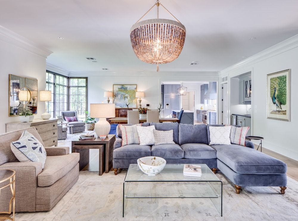 A bright, elegant living room features a blue sectional sofa, a beige armchair, a glass coffee table, and a beaded chandelier.