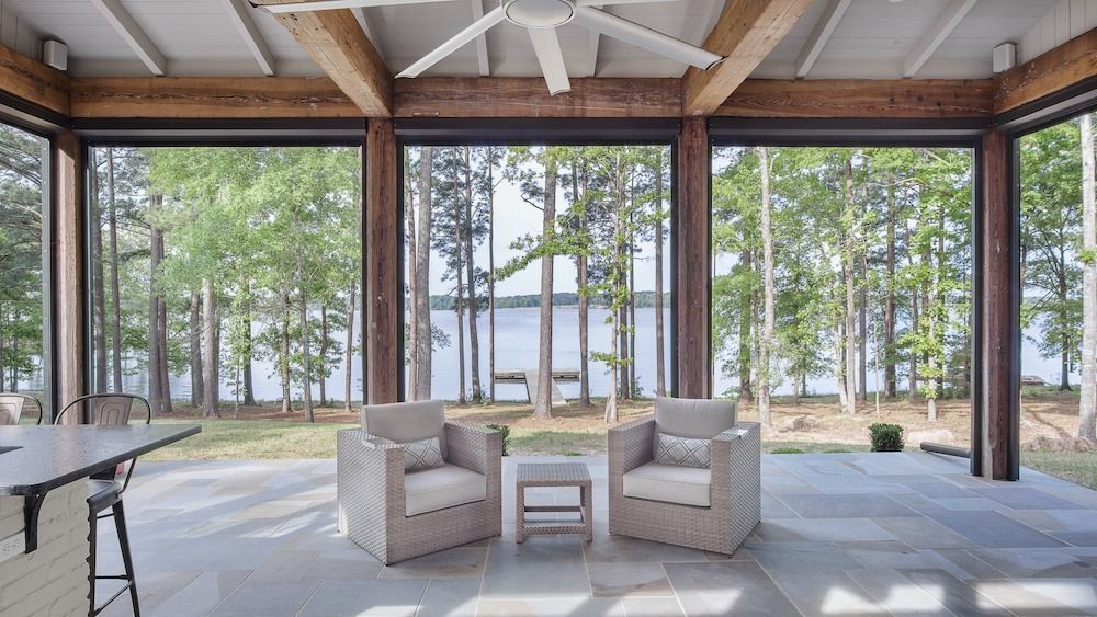 Two beige armchairs and a small table sit on a covered stone patio overlooking a lake and forest.