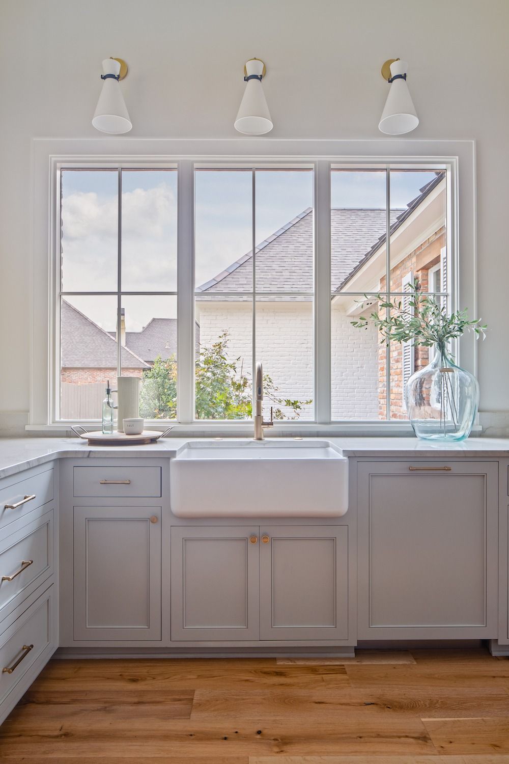 Kitchen sink area featuring a white farmhouse sink, grey cabinetry, hardwood floors, and a window with three light fixtures.