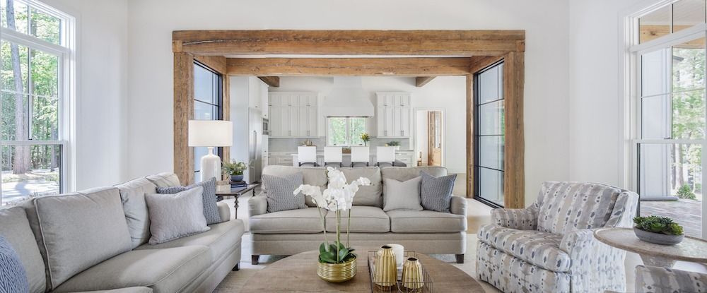 Living room with light gray couches and a patterned armchair, featuring a wooden beam doorway to a kitchen background.
