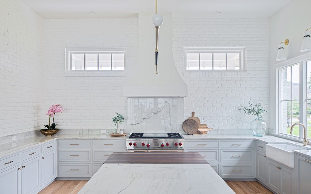 A bright, modern kitchen featuring white brick walls, light gray cabinetry, a marble-topped island, and a range stove.