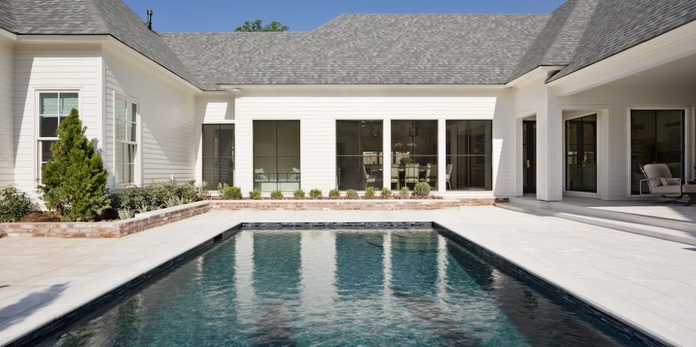 A rectangular swimming pool sits in front of a white house with large glass windows and gray roof shingles.