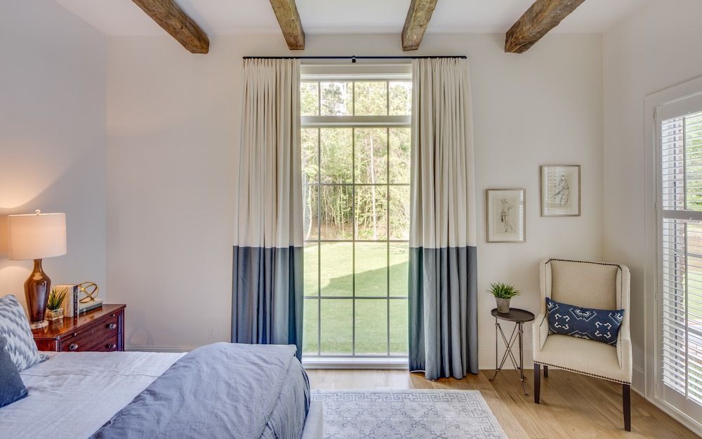 Bedroom with a bed, a window with two-toned beige and blue curtains, a patterned armchair, and exposed ceiling beams.