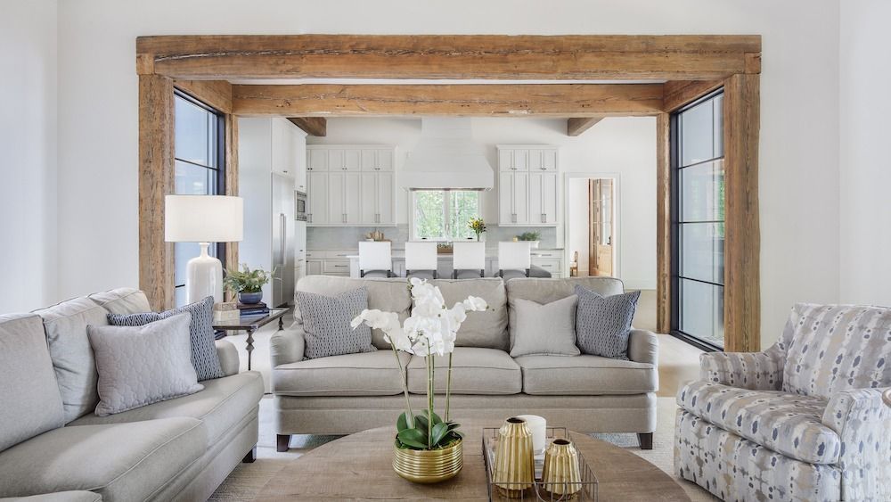 A living room with light gray sofas, a patterned armchair, and a wood-beamed doorway leading to an open-plan kitchen.