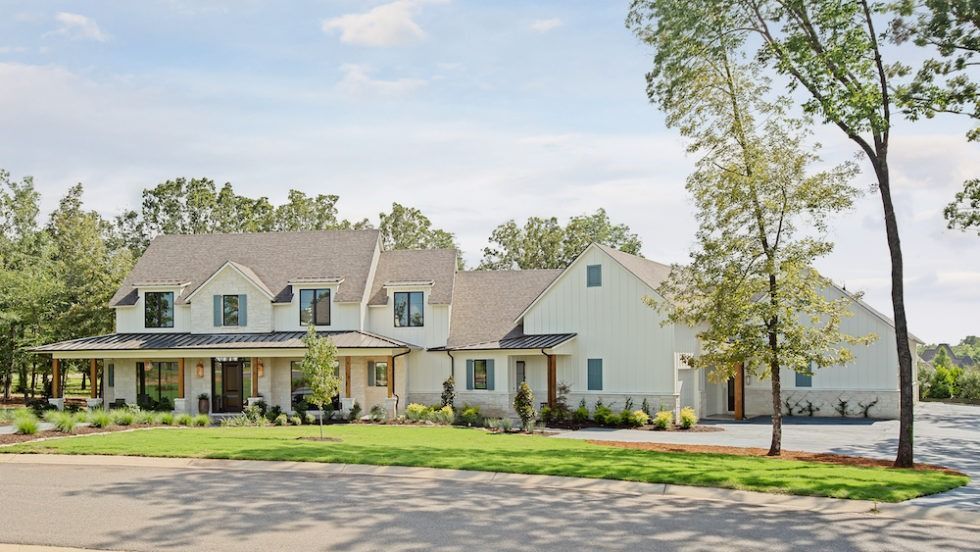 A sprawling white farmhouse with a dark roof and covered porch, set on a manicured lawn surrounded by mature trees.