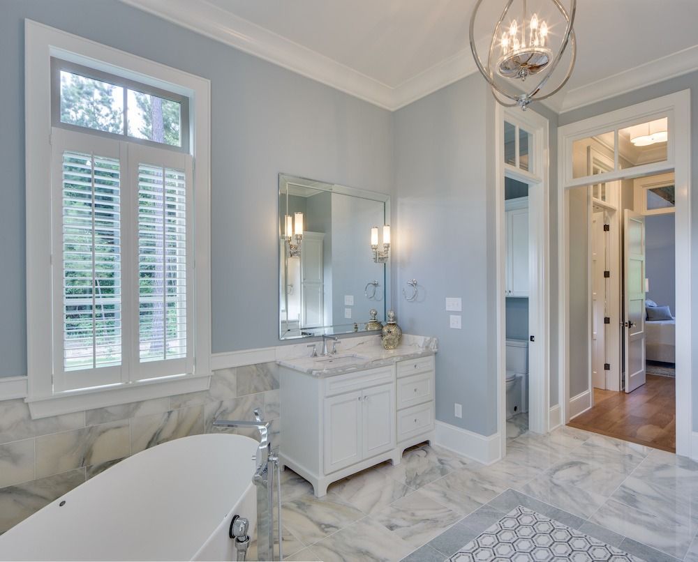 A bright, elegant bathroom with a white freestanding tub, marble floors, a vanity with a mirror, and blue-gray walls.