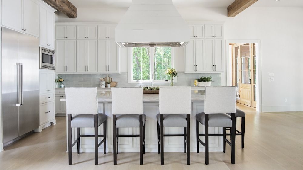 A white modern kitchen featuring a central island with four bar stools, a large range hood, and stainless steel fridge.