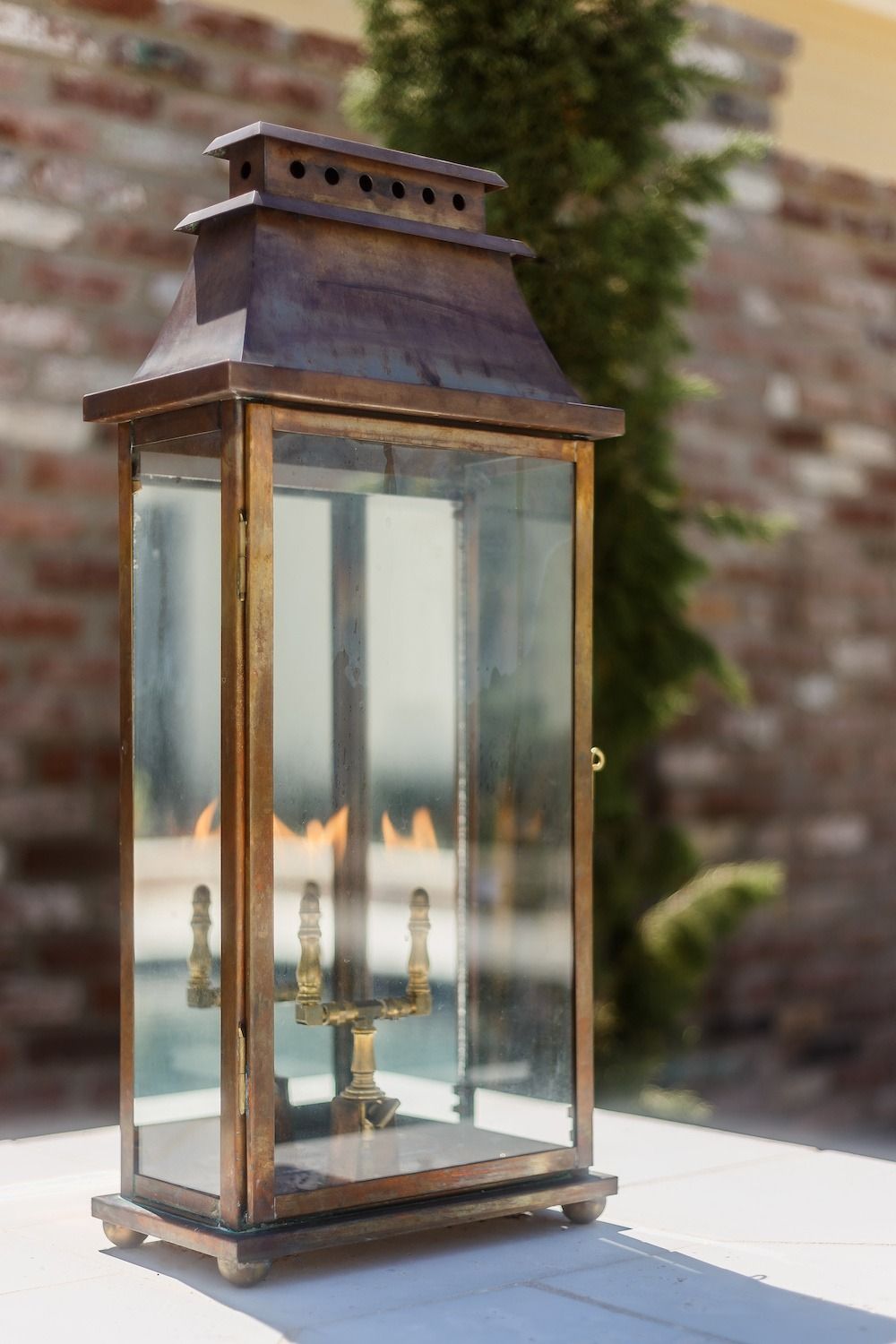 A brass lantern with lit flames inside sits on a white surface, set against a blurred brick wall and evergreen tree.