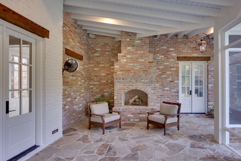 A covered patio with a brick fireplace, two beige armchairs, white framed doors, and a stone floor.