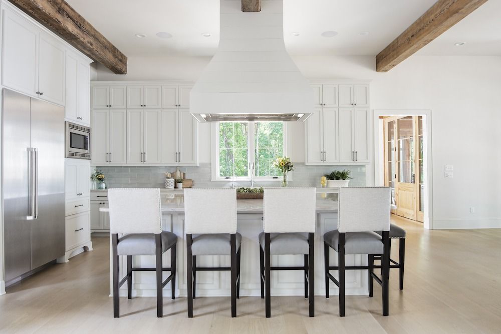 A modern white kitchen with a large central island, four bar stools, wood ceiling beams, and stainless steel appliances.